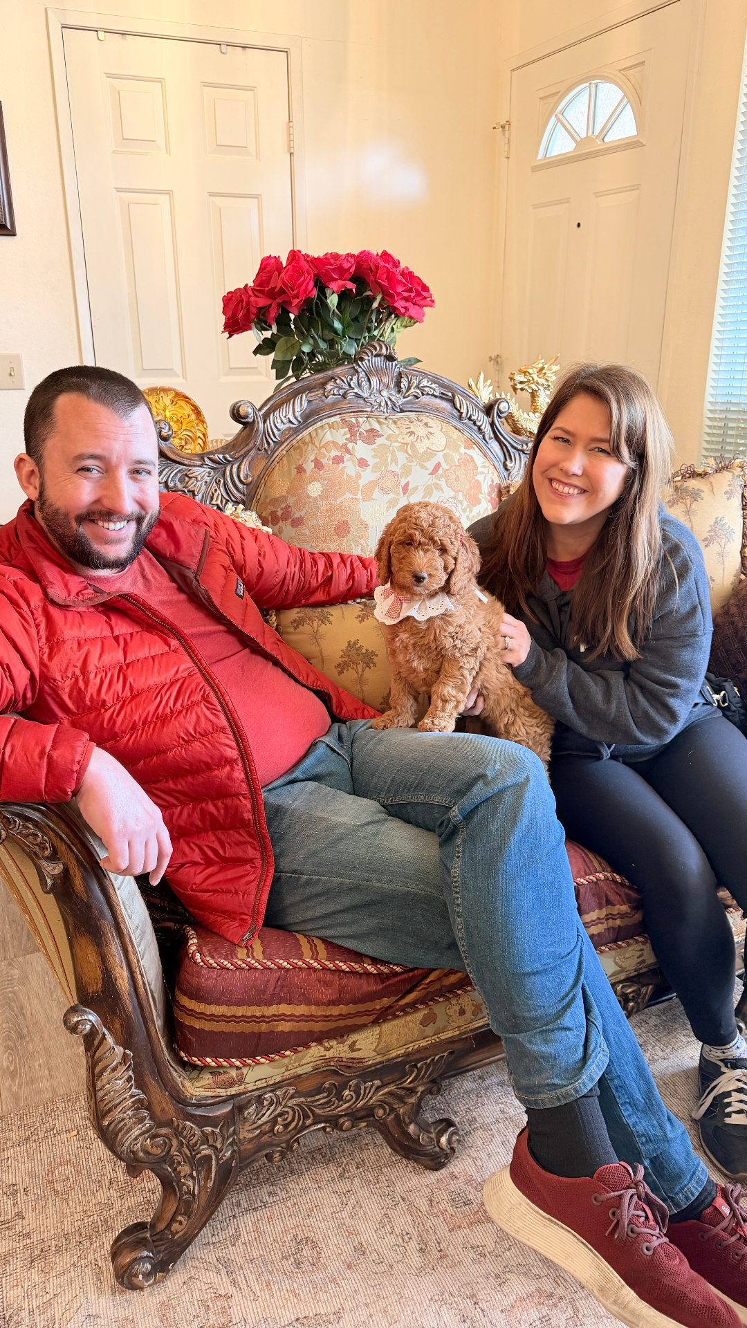 A Kid's Best Doodle homecoming — a couple in a red jacket seated together with their mini Goldendoodle puppy on an ornate chair