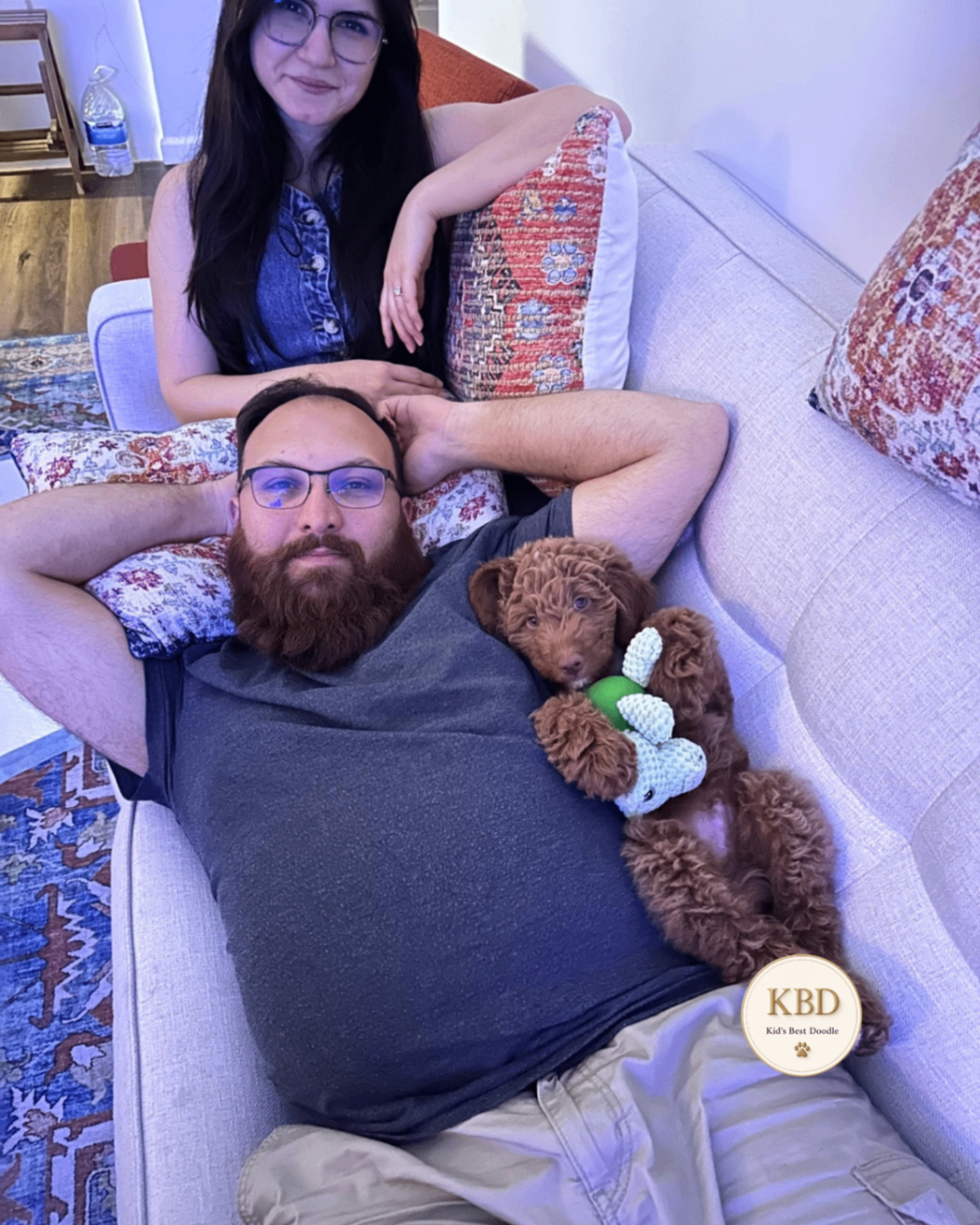 A couple on the couch with their Goldendoodle puppy peeking over — Kid's Best Doodle California